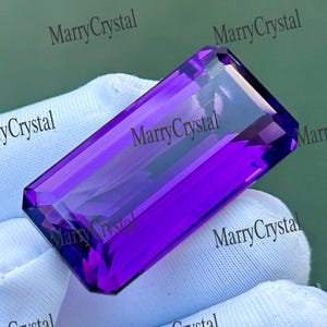 Può includere: Una gemma di ametista rettangolare sfaccettata con una tonalità viola intenso e una superficie lucida. La pietra presenta bordi netti e una qualità trasparente, mostrando un taglio brillante che riflette la luce sulle sue sfaccettature.