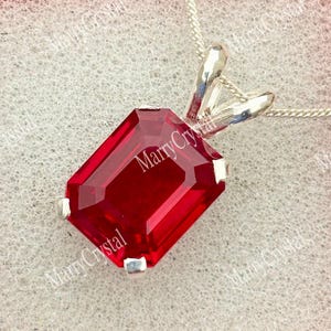 Op de afbeelding: Een rechthoekige, gefacetteerde, dieprode edelsteen hanger in een zilverkleurige metalen zetting. De hanger hangt aan een fijne zilverkleurige ketting en wordt gepresenteerd in een vierkant, lichtroze doosje met een witte schuimrubberen inzet.