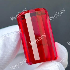 May include: A rectangular, faceted gemstone with a vibrant, translucent red color. The stone features sharp, clean edges and a polished surface that reflects light, creating distinct vertical highlights across its deep crimson, glass-like body.