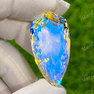May include: A large, faceted crystal pendant with a teardrop shape. The crystal has a vibrant blue hue in the center, transitioning to yellow and iridescent colors at the edges. The crystal is held by a gloved hand against a green background.