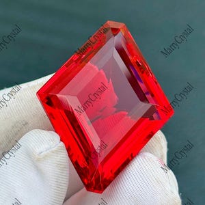 May include: A vibrant, square-cut red gemstone with sharp, faceted edges that catch the light. The stone displays a deep, saturated crimson hue and a high-gloss, transparent finish, showcasing precise geometric cuts across its surface.
