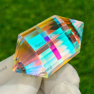 May include: A clear, prism-cut crystal with iridescent rainbow hues. The crystal is held in a gloved hand against a green background. The text "MarryCrystal" is visible on the image.