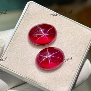 May include: Two oval-shaped, deep red gemstones with a star-like pattern are displayed in a white box. The gems are held by a gloved hand. The gems have a polished, reflective surface.