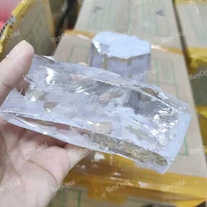 Puede incluir: Un espécimen de cristal rectangular, claro y translúcido, con una textura superficial rugosa e irregular. El mineral muestra fracturas internas e inclusiones blancas nubladas en toda su estructura, reflejando la luz en sus bordes facetados.