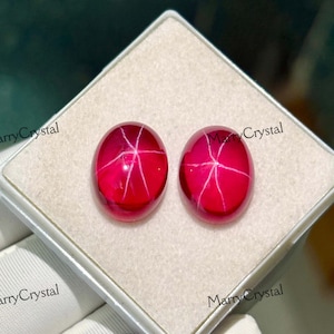 Peut inclure: Deux pierres précieuses ovales rouge vif avec un motif en étoile, présentées dans une boîte blanche. Les pierres précieuses ont une finition polie et brillante. Le texte "MarryCrystal" est visible en arrière-plan. Idéal pour la joaillerie.