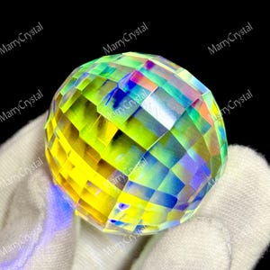 May include: A faceted crystal sphere with a vibrant display of colors, including yellow, green, blue, and pink. The sphere is held in a white glove, with the text "MarryCrystal" visible.