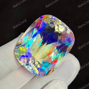 May include: A large, iridescent gemstone with a rectangular cushion cut. The gem displays a spectrum of colors, including blues, greens, yellows, and oranges, reflecting light. The stone is held by a gloved hand. The text "MarryCrystal" is repeated around the image.