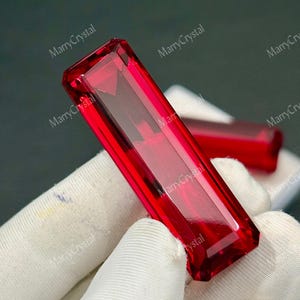 May include: A faceted, rectangular red gemstone. The gem is held by a gloved hand, with another gem visible in the background. The gem has a polished, reflective surface. The text "MarryCrystal" is visible in the background.