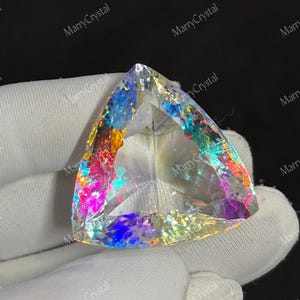 May include: A large, triangular gemstone with iridescent rainbow colors. The crystal is held in a white glove, showcasing its facets and light refraction. The stone's edges are clearly defined, and the overall effect is striking.