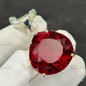 May include: A large, faceted, oval-shaped, deep red gemstone pendant. The gemstone is set in a silver-colored metal setting with a decorative bail. The pendant is held by a gloved hand.