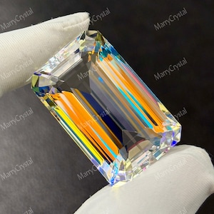 May include: A large, rectangular gemstone with an emerald cut. The crystal is held by a gloved hand. The gem refracts light, displaying a spectrum of colors including orange, blue, and yellow. The background is dark.