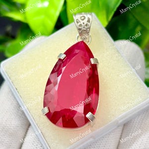 May include: A teardrop-shaped, faceted, red gemstone pendant. The pendant is set in a silver-colored metal with a decorative bail. The gemstone is displayed in a white box, with a green leafy background.