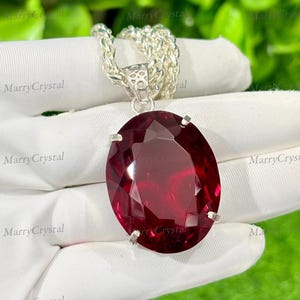 May include: A large, oval-cut, deep red gemstone pendant on a silver chain. The gemstone is faceted, catching the light. The chain is a thick, textured silver. The pendant is held by silver prongs.