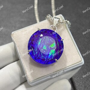 May include: A round, faceted pendant featuring a deep blue and iridescent green color palette, set in a silver-toned metal prong setting. The pendant hangs from a silver-toned chain and is displayed on a square, light-colored jewelry box.