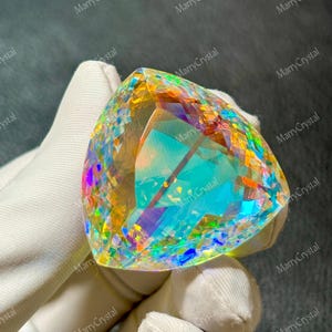 May include: A large, triangular-cut gemstone with vibrant, iridescent colors. The gem displays a spectrum of hues, including blues, greens, yellows, and purples, with internal facets creating a dazzling effect. The gem is held by a gloved hand.