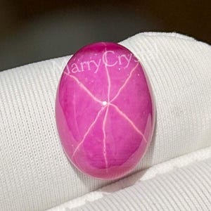 May include: A polished, oval-shaped, pink gemstone with a six-rayed star pattern. The gem is a vibrant magenta color and appears to be set against a white background. The gem is approximately 1.5 inches long.