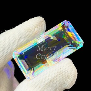 May include: A rectangular, iridescent crystal with the text "Marry Crystal" etched on its surface. The crystal displays a spectrum of colors, including blues, greens, and yellows, and is held by a gloved hand against a black background.
