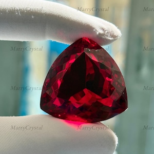 May include: A faceted, triangular-shaped, deep red gemstone. The gem is held by a gloved hand, showcasing its brilliant cut and vibrant color. The gem's facets reflect light, creating a sparkling effect. The overall theme is jewelry and gemstones.