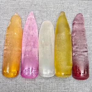 Puede incluir: Cinco objetos cilíndricos, semitranslúcidos y alargados con puntas redondeadas, dispuestos verticalmente. Los colores de izquierda a derecha son ámbar, rosa claro, transparente, amarillo y morado oscuro. Cada pieza tiene una superficie lisa y texturizada.