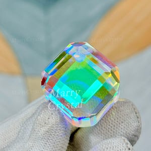 May include: A faceted, iridescent crystal in a square shape. The crystal reflects a spectrum of colors, including green, blue, and yellow. The text "Marry Crystal" is visible on the crystal. The crystal is held by a gloved hand.