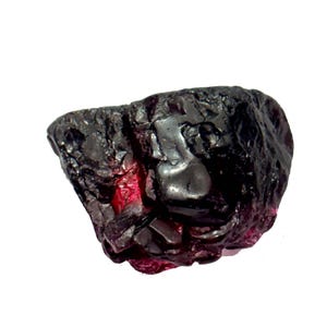 Puede incluir: Una gema en bruto, sin cortar, con un exterior oscuro, casi negro. La piedra tiene una superficie texturizada con destellos de rojo intenso y púrpura. La forma irregular resalta su forma natural y cruda.