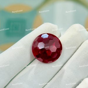May include: A faceted, round, deep red gemstone. The gem is held in a white gloved hand. The gem has a multi-faceted surface that reflects light. The text "MarryCrystal" is visible in the background.