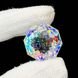 May include: A multi-faceted, clear crystal with rainbow reflections. The crystal is held by a white fabric. The text "Marry Crystal" is visible on the crystal's surface. The crystal has a geometric shape.