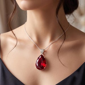 May include: A teardrop-shaped, deep red faceted gemstone pendant suspended from a delicate silver chain. The pendant features a polished silver setting with intricate metalwork at the top, securing the large, vibrant red stone in a classic prong style.