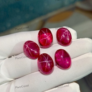 Peut inclure: Cinq pierres précieuses ovales rouge foncé avec un effet étoile. Les gemmes ont une surface polie et brillante et un motif d'étoile blanc sur leur face. Les gemmes sont présentées sur une surface blanche. Le texte "MarryCrystal" est visible.