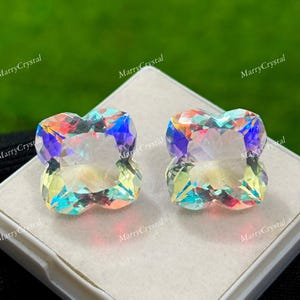 May include: Two iridescent, multi-colored gemstones with a four-leaf clover shape. The gems are displayed on a white surface, with the text "MarryCrystal" repeated around the edges. The background is a blurred green.