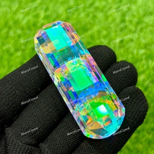 May include: A rectangular, iridescent crystal with multiple facets, reflecting a spectrum of colors including green, blue, and yellow. The crystal is held against a black surface, with the text "MarryCrystal" repeated around the edges.