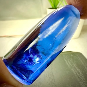 May include: A smooth, cylindrical glass object with a deep, translucent blue color. The surface features subtle, fine horizontal striations throughout the length of the piece, creating a reflective texture that catches light across its curved form.