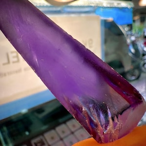 May include: A translucent, elongated purple crystal shard with a smooth, polished surface and a jagged, textured base. The deep violet hue transitions to lighter shades, revealing internal fractures and natural variations within the stone structure.