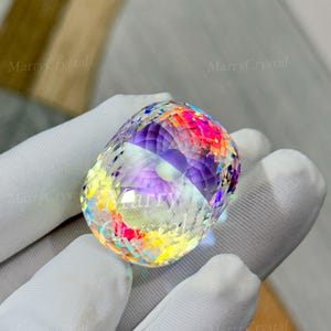 May include: A faceted, oval-shaped crystal with a rainbow of colors, including pink, yellow, and purple. The crystal is held in a white gloved hand. The text "MarryCrystal" is visible on the crystal.
