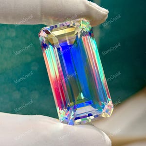May include: A large, rectangular crystal with an emerald cut, held by a gloved hand. The crystal refracts light, displaying a spectrum of colors including blue, red, and yellow. The text "MarryCrystal" is visible.