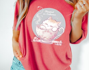 May include: Red-orange t-shirt featuring a graphic of a cat in a teacup with the text "Steeped in Cuteness". The cat is white and orange, and the teacup has a floral design. The shirt is short-sleeved.