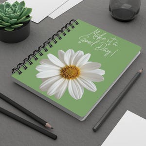 May include: A spiral-bound notebook with a sage green cover featuring a large white daisy with a yellow center. The text "Make it a Good Day!" is printed in white script in the upper right corner. The notebook has black wire binding.