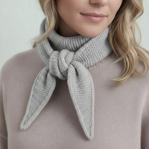 Puede incluir: Bufanda de punto gris claro anudada al cuello. La bufanda está hecha de un hilo suave y texturizado. Los extremos de la bufanda cuelgan en forma triangular. La bufanda es de color neutro y se puede usar con muchos atuendos. Accesorio versátil.