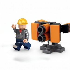 K&ouml;nnte beinhalten: Eine Spielfigur aus Klemmbausteinen mit gelbem Bauhelm, grauem Kapuzenpullover &uuml;ber einem roten T-Shirt und dunkelblauer Hose, neben einem kleinen, aus Bausteinen zusammengesetzten mechanischen Bauger&auml;t in Orange und Schwarz.
