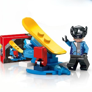 K&ouml;nnte beinhalten: Eine Kunststoff-Spielfigur mit schwarzem Helm und blauer Jacke mit R-Emblem, die neben einer blau-roten Konstruktionsbasis steht, welche ein gro&szlig;es gelbes snowboardartiges Teil und eine rote Schachtel mit Produktabbildungen enth&auml;lt.