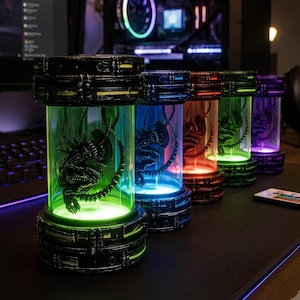 Op de afbeelding: Een rij van vijf cilindrische lampen, elk met een donker, gedetailleerd xenomorph-wezenfiguur. De lampen hebben zwarte, getextureerde basissen en bovenkanten met verlichte transparante centra in de kleuren groen, blauw, oranje, groen en paars.