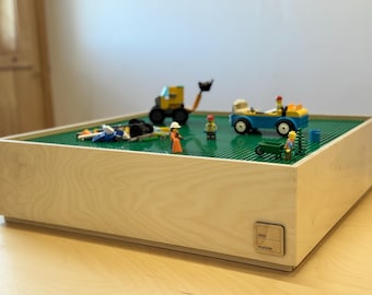 May include: A wooden toy construction table with a green base, featuring toy construction vehicles and figures. The table is rectangular and has a light wood finish. The table measures approximately 46 cm wide and 46 cm long.