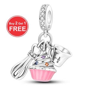 May include: Silver charm with a cupcake, whisk, and measuring cup design. The cupcake has a pink base and white frosting with colorful gems. The charm has a ring with clear stones and the text "Buy 2 Get 1 FREE" in a red circle.