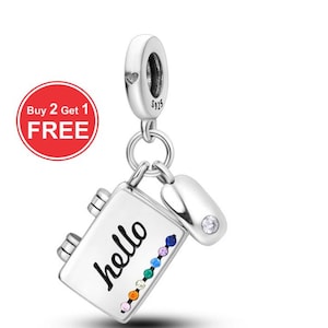 Puede incluir: Un charm de plata con un diseño de cuaderno que dice "hello" en escritura negra, acentuado con gemas de colores. Un pequeño charm a juego con una sola gema transparente cuelga junto al cuaderno. El charm está sujeto a un lazo plateado.