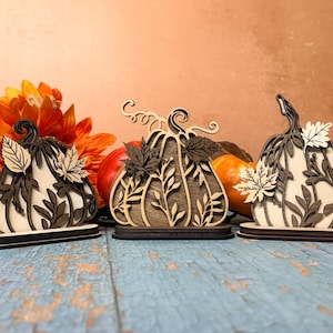 May include: Three decorative wooden pumpkin ornaments standing on individual bases. Each pumpkin features intricate laser-cut details including layered leaf patterns and swirling stems, crafted from light and dark wood tones for a layered effect.