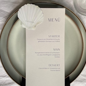 May include: A rectangular white menu card with the text MENÜ, STARTER, MAIN, and DESSERT, featuring a white seashell decoration with the name KATERINA printed on it. The menu is placed on a light gray plate with a dark rim and a black napkin.
