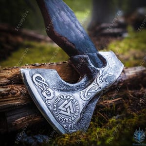 May include: A silver axe head with intricate engraved designs and a dark wooden handle. The axe head features a circular emblem with runes and a geometric symbol. The handle is dark and appears to be made of wood. The axe is resting on a piece of wood.