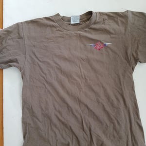 May include: A brown t-shirt with a crew neck and short sleeves. The shirt features a small graphic on the chest, a stylized rose with red, pink, and blue accents. The shirt is made of a soft, slightly wrinkled fabric.
