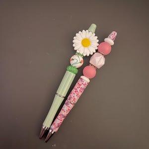 May include: Two decorative pens. One pen is light green with a white daisy and green and white beads. The other pen is pink with a floral pattern and pink beads. Both pens have silver accents.