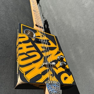 May include: A handmade cigar box guitar with a black and yellow design. The guitar features a wooden neck, strings, and tuning pegs. The body of the guitar has a circular sound hole and a metal tailpiece. The guitar is angled towards the viewer.